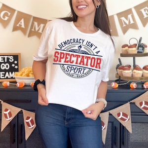 Può includere: T-shirt bianca con la scritta "DEMOCRACY ISN'T A SPECTATOR SPORT" in rosso e blu. La maglietta ha girocollo e maniche corte. Il design include una torcia e una corona d'alloro.