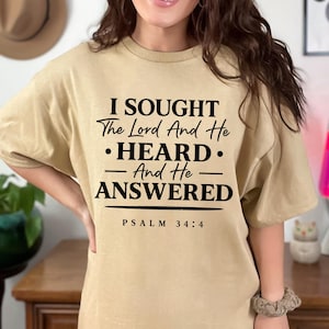 Peut inclure: T-shirt beige avec l'inscription noire : "I SOUGHT The Lord And He • HEARD • - And He - ANSWERED PSALM 34:4". Le t-shirt a des manches courtes et une coupe ample.