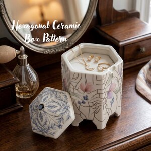 Può includere: Una scatola esagonale in ceramica con finitura smaltata a crepa, caratterizzata da motivi floreali dipinti in rosa e viola. Il coperchio è decorato con motivi a foglie blu. All'interno sono visibili una collana a mezzaluna dorata, due piccoli orecchini e un anello.
