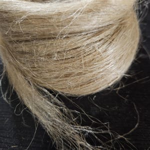 Può includere: Primo piano di una palla di spago color naturale. Lo spago sembra essere fatto di un materiale fibroso, con una consistenza leggermente ruvida e una tonalità marrone chiaro. Fili di spago si estendono verso l'esterno dalla palla.