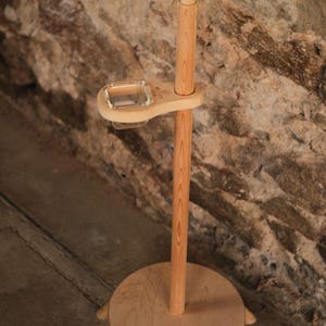Könnte beinhalten: Ein hoher Holzständer mit einem runden Fuß und einem vertikalen Stab. Am Stab ist eine kleine, rechteckige Glasschale in einem Holzrahmen befestigt. Die Spitze des Stabes ist mit einem Büschel aus roten und beigen Fasern verziert.