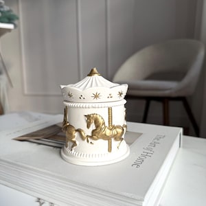 May include: A white ceramic carousel-themed trinket box with gold accents. The box features a carousel design with gold horses and poles. The lid is white with gold stars and a gold finial. The box is sitting on a book.