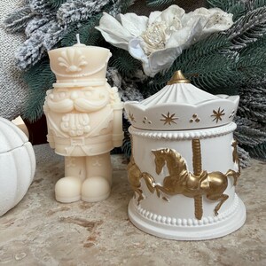 Può includere: Una candela beige a forma di schiaccianoci e un barattolo bianco a tema giostra con dettagli dorati. Il barattolo della giostra presenta cavalli dorati e dettagli a stella. La candela ha la forma di un soldato schiaccianoci.