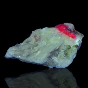 May include: A close-up of a mineral specimen with a rough, textured surface. The stone is primarily off-white with areas of bright red and hints of green. The background is black, and the mineral's reflection is visible.