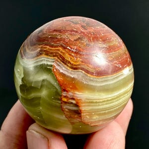 Op de afbeelding: Een gepolijste onyx bol met banden van groen, rood en bruin. De edelsteenbal heeft een glad, reflecterend oppervlak en een natuurlijk, aards uiterlijk. De bol is een decoratief item.