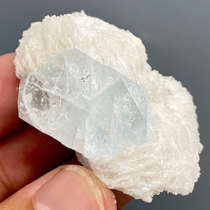 May include: A clear, light blue crystal formation with a geometric shape is set against a backdrop of white, textured mineral. The crystal is held in a hand, showcasing its facets and clarity. The overall composition highlights the natural beauty of the mineral specimen.