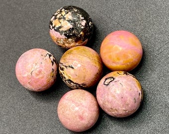 Könnte beinhalten: Sechs polierte Edelstein-Kugeln in Rosa-, Pfirsich- und Schwarztönen. Die Kugeln haben eine glatte, abgerundete Form und unterschiedliche Muster. Die Edelsteine sind auf einer dunkelgrauen Oberfläche angeordnet.