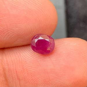 May include: A faceted, oval-shaped ruby gemstone. The gem is a deep, saturated red color with multiple facets that reflect light. The ruby is held between two fingers, showcasing its size and brilliance.