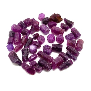 May include: A collection of raw, unpolished ruby crystals in various shades of deep magenta, crimson, and dark purple. These small, naturally faceted stones exhibit irregular shapes and textures, showcasing a mix of translucent and opaque surfaces.