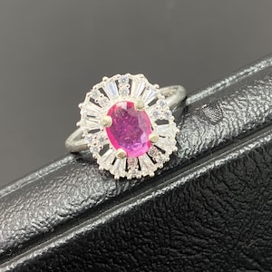 May include: A silver ring featuring a large, oval-cut, magenta gemstone at the center, surrounded by a halo of smaller, clear stones and baguette-cut accents. The ring is set against a dark background.
