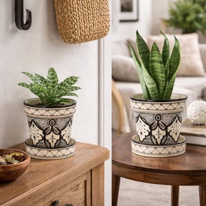 Op de afbeelding: Twee decoratieve keramische plantenpotten met bijpassende schotels. De potten zijn crèmekleurig met een zwart geometrisch patroon. Eén pot bevat een plant met groen-witte gevlekte bladeren, de andere een sansevieria.