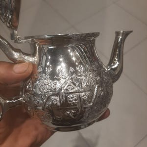 Peut inclure: Une théière en argent avec des motifs floraux et géométriques en relief. La théière a un bec verseur incurvé, un couvercle à charnière et une anse. Le métal poli reflète la lumière, soulignant le design détaillé. La théière est tenue dans une main.