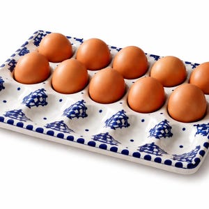 Può includere: Un portauova in ceramica con dieci uova marroni. Il vassoio bianco presenta un motivo floreale blu e un bordo a pois. Le uova sono disposte in scomparti individuali.