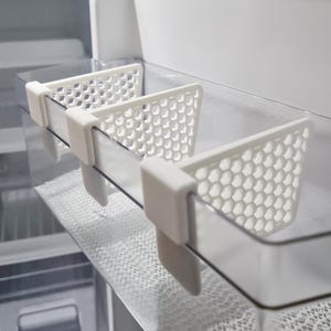 Könnte beinhalten: Weiße Kunststoff-Kühlschrank-Regalteiler mit Wabenmuster. Die Teiler sind so konzipiert, dass sie an der Ablage befestigt werden und separate Fächer schaffen. Das Bild zeigt die Teiler, die in einem transparenten Kunststoffregal installiert sind.