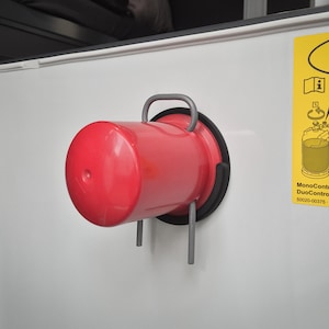 Può includere: Un serbatoio di propano rosso con una maniglia nera e un gancio argentato è montato su una parete bianca. Il serbatoio è etichettato "MonoControl CS DuoControl CS 500020-00375 - 04 - 05/2021 ©".