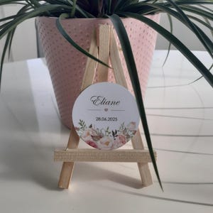 Könnte beinhalten: Ein kleiner Holzstaffelei mit einem runden weißen Schild, auf dem "Eliane" mit einem kleinen rosa Herz und dem Datum "28.06.2025" steht. Das Schild hat ein florales Design mit rosa und weißen Blumen.