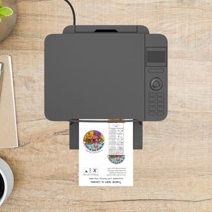 May include: A gray printer is printing a document with colorful graphics and text. The document features a circular design with various images and the text "Twelve Spies in Canaan". The printer sits on a wooden surface.