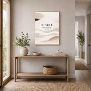 Può includere: Una stampa artistica incorniciata con le parole "BE STILL and know that I am God" in una palette di colori neutri. L'opera d'arte è appesa sopra una consolle in legno con un cestino intrecciato, una ciotola di ceramica e un vaso con verde. Un tappeto neutro è di fronte al tavolo.
