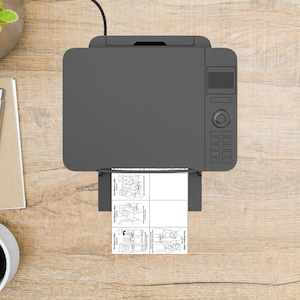 May include: A grey printer on a wooden surface, printing a black and white document with illustrations and text. A pen, notepad, and coffee cup are also visible. The printer is connected to a power cable.