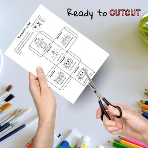 May include: A white paper with a Kindness Cube design is being cut with scissors. The cube features illustrations and phrases like "Smile," "Thank You," and "Share Toys." The words "Ready to CUTOUT" are written at the top.