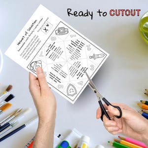 May include: A white paper craft activity titled "Helmet of Salvation" is being cut with scissors. The text "Ready to CUTOUT" is above the paper. Paintbrushes, paint tubes, and colored pencils are in the background.