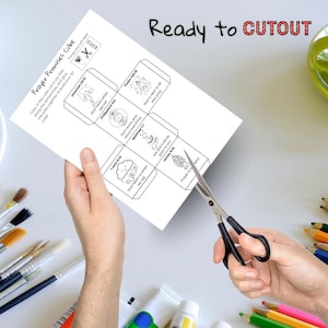 May include: A white paper craft titled "Prayer Promises Cube" with illustrations and Bible verses. The paper is being cut with scissors. Text on the paper includes "Ready to CUTOUT".