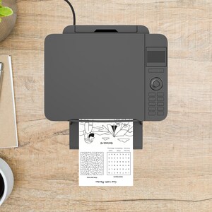 May include: A top-down view of a gray printer printing a black and white activity sheet. The sheet includes a maze, word search, and the text "Genesis 12". The printer sits on a light brown wooden surface.
