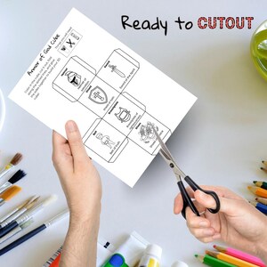 May include: A white paper with the text "Armor of God Cube" and illustrations of a helmet, shield, sword, breastplate, belt, and shoes. The words "Ready to CUTOUT" are written at the top. Scissors are cutting the paper.