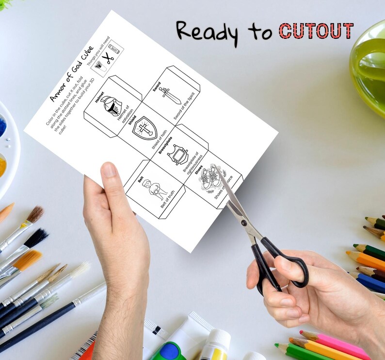 May include: A white paper with the text "Armor of God Cube" and illustrations of a helmet, shield, sword, breastplate, belt, and shoes. The words "Ready to CUTOUT" are written at the top. Scissors are cutting the paper.