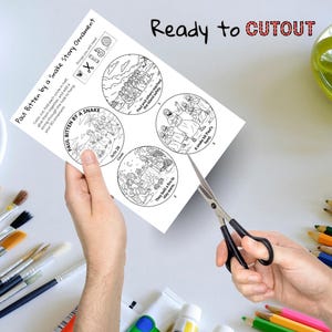 May include: A black and white coloring sheet with the text "Paul Bitten by a Snake Story Ornament" and illustrations. The sheet is being cut with scissors. The words "Ready to CUTOUT" are at the top.