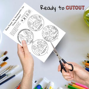 May include: A white paper with a black and white illustration of a Bible story, titled "Elisha Raises the Shunammite's Son Story Ornament." The text "Ready to CUTOUT" is at the top. Scissors are cutting the paper.