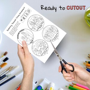 May include: A sheet of paper with a "Doubting Thomas Story Ornament" design, featuring illustrations and text. The words "Ready to CUTOUT" are written at the top. Scissors are cutting the paper. Paintbrushes and colored pencils are in the background.