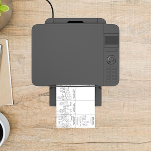 Puede incluir: Una impresora gris est&aacute; imprimiendo una imagen en blanco y negro con cuatro paneles. La impresora est&aacute; sobre una superficie de madera marr&oacute;n claro. Se ven un bol&iacute;grafo, un bloc de notas y una taza de caf&eacute;.