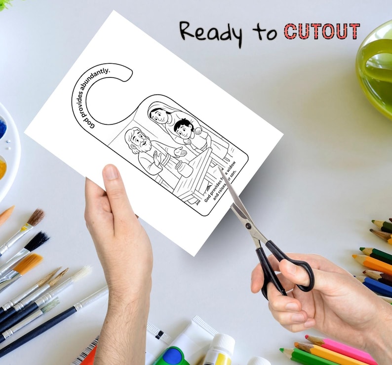 Op de afbeelding: Een zwart-witte deurhanger met een bijbelse illustratie van een gezin. De tekst "God voorziet overvloedig" en "Ready to CUTOUT" zijn zichtbaar. Een schaar knipt de deurhanger.