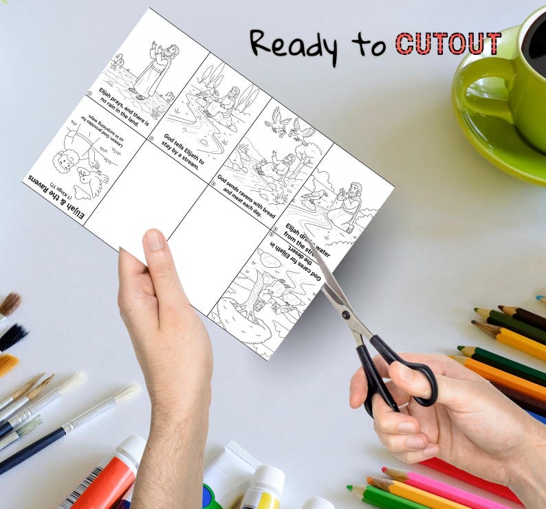 May include: A person is cutting out a black and white illustrated children's activity sheet with scissors. The sheet features Bible story panels with text: "Elijah & the Ravens" and "Ready to CUTOUT". A green mug and colored pencils are in the background.