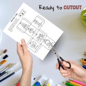 May include: A white paper with a "Worship Cube" design is being cut with scissors. The cube features illustrations and phrases like "Sing," "Clap your hands in joy," and "Dance before the Lord." The words "Ready to CUTOUT" are written at the top.