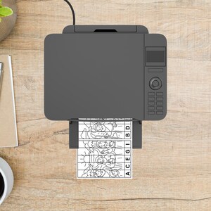 May include: A gray printer is printing a black and white image of a puzzle on a wooden desk. The puzzle shows a line drawing of people and the letters ACEGIBD. A pen, notepad, and coffee cup are also on the desk.