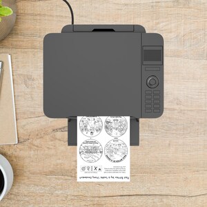 May include: A gray inkjet printer is printing a sheet of paper with four circular illustrations and text. The text reads "Paul Bitten by a Snake Story Ornament". The printer is on a light brown wooden surface.