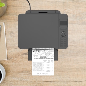 May include: A gray printer is shown from above, printing a black and white activity sheet. The sheet includes a maze, word search, and an illustration with the text "Moses strikes the rock, and water pours out for the people (Numbers 20)."