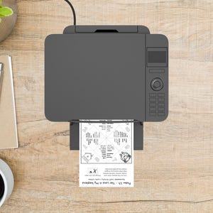 May include: Overhead view of a gray printer printing a black and white document. The document has a circular design with text and a sheep illustration. The printer is on a wooden surface, with a pen, notepad, and coffee cup visible.