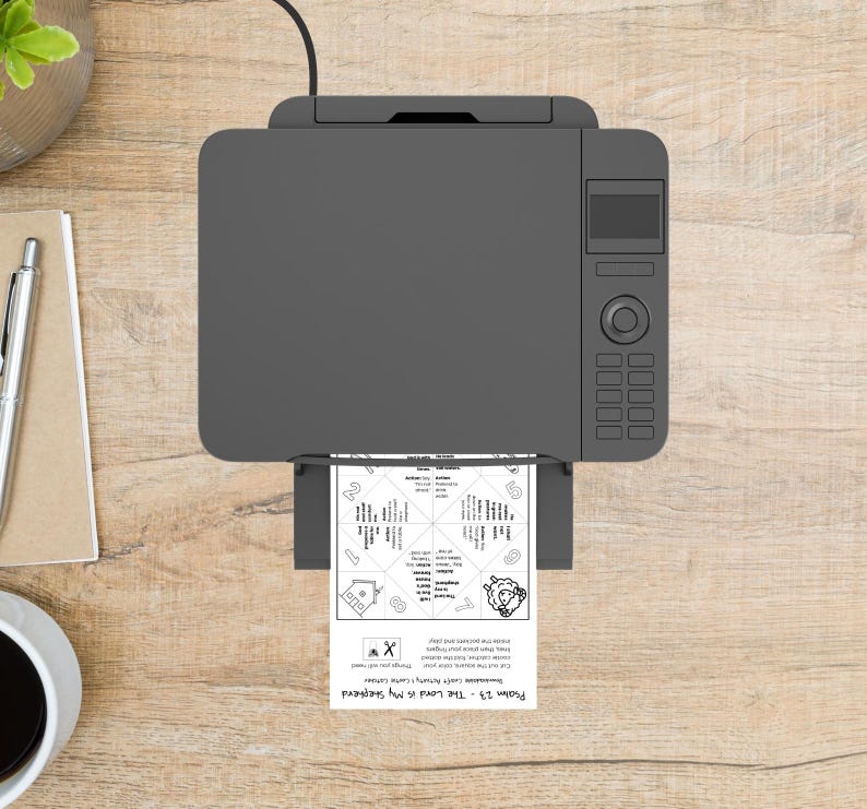 May include: Overhead view of a gray printer printing a black and white document. The document has a circular design with text and a sheep illustration. The printer is on a wooden surface, with a pen, notepad, and coffee cup visible.