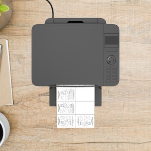 May include: A gray printer is printing four small black and white images. The images have text and illustrations. The printer is on a light brown wooden surface. A pen, notepad, and coffee cup are visible.