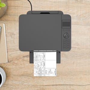 May include: Overhead view of a gray printer with a sheet of paper emerging from the output tray. The paper has four black and white illustrations and the text "The Lost Sheep". The printer is on a light brown wooden surface.