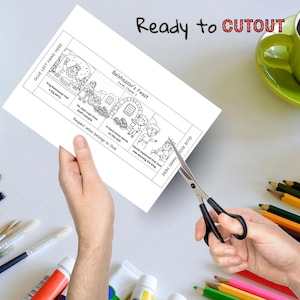 May include: A black and white illustrated paper with the text "Belshazzar's Feast" and "Ready to CUTOUT". The paper is being cut with scissors. A green mug and coloured pencils are in the background.