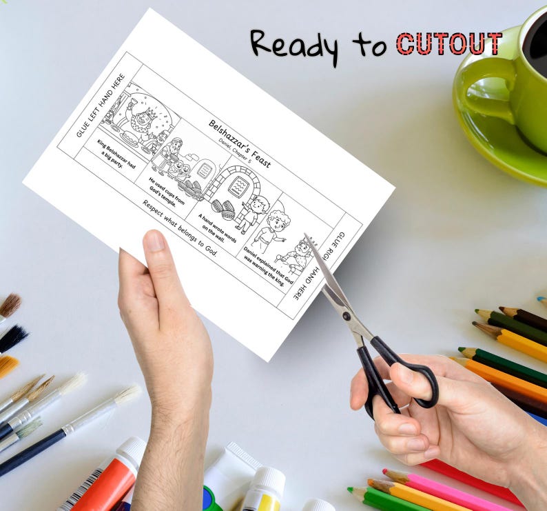 May include: A black and white illustrated paper with the text "Belshazzar's Feast" and "Ready to CUTOUT". The paper is being cut with scissors. A green mug and coloured pencils are in the background.