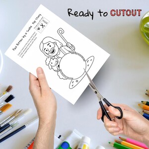 May include: A black and white coloring page titled "Paul Bitten by a Snake Flip Story" is being cut with scissors. The page features a cartoon illustration of a man with a sheep and the text "Ready to CUTOUT". Paintbrushes and colored pencils are in the background.