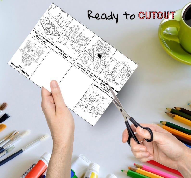 May include: A black and white illustrated cutout sheet with Bible story panels, including text like "The Paralyzed Man" and "Ready to CUTOUT." The sheet is being cut with scissors. Colored pencils and a green mug are in the background.
