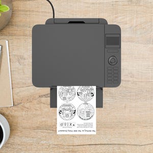 May include: A gray printer printing a black and white circular design on paper. The design features illustrations and text, including the words "The Writing on the Wall Story Ornament."