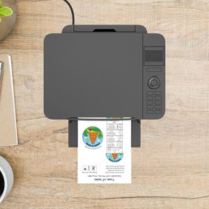 May include: A gray printer printing a sheet of paper with a colorful graphic of a tower and the text "Tower of Babel". The printer is on a light brown wooden surface, with a pen, notepad, and coffee cup visible.