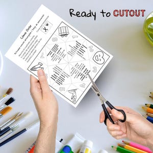May include: A white paper with the text "C&oacute;mo Orar" and "Ready to CUTOUT" is being cut with scissors. The paper has a geometric design with numbers and illustrations. Paintbrushes and colored pencils are in the background.